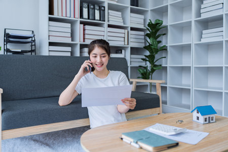 Young woman engaged in a phone conversation while working on home finances in a cozy modern living room.の写真素材