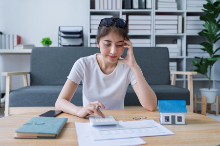 Young woman analyzing financial documents at home with calculator and model house on table in modern living room.の写真素材