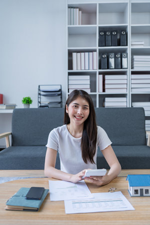 Young woman analyzing financial documents at home with calculator and model house on table in modern living room.の写真素材