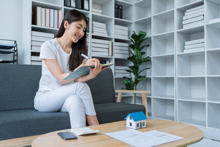 Young woman analyzing financial documents at home with calculator and model house on table in modern living room.の写真素材