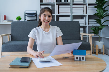 Young woman analyzing financial documents at home with calculator and model house on table in modern living room.の写真素材