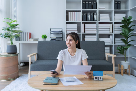 Young woman engaged in a phone conversation while working on home finances in a cozy modern living room.の写真素材