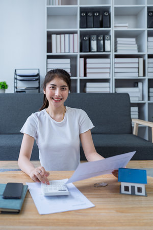 Young woman analyzing financial documents at home with calculator and model house on table in modern living room.の写真素材