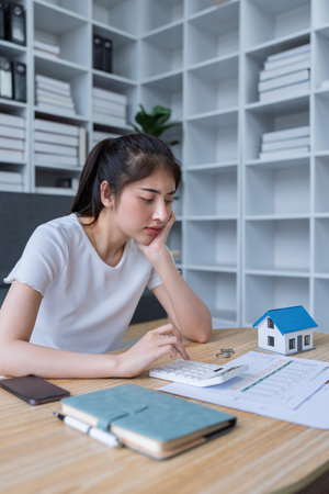 Young woman analyzing financial documents at home with calculator and model house on table in modern living room.の写真素材