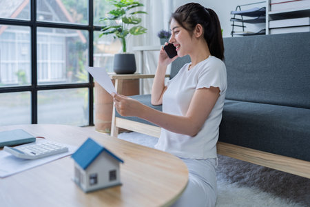 Young woman engaged in a phone conversation while working on home finances in a cozy modern living room.の写真素材