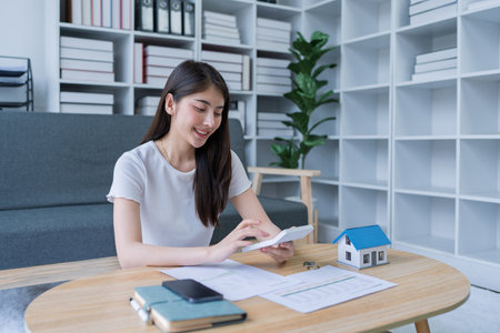 Young woman analyzing financial documents at home with calculator and model house on table in modern living room.の写真素材