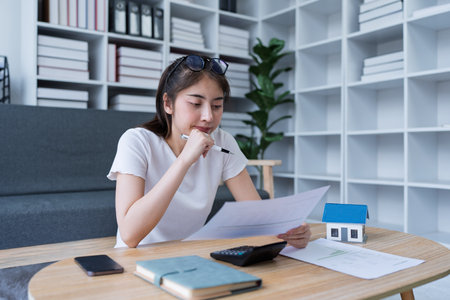 Thoughtful woman reviewing documents while calculating expenses for real estate investment at home office setting.の写真素材