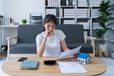 Thoughtful woman reviewing documents while calculating expenses for real estate investment at home office setting.の写真素材