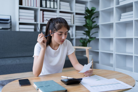 Young woman analyzing receipt and financial documents at home office while experiencing confusion and concern.の写真素材