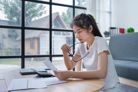 Young woman analyzing receipt and financial documents at home office while experiencing confusion and concern.の写真素材