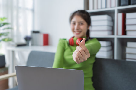 Woman with make mini heart symbol, wearing headphones using her computer enjoying music at home.の写真素材
