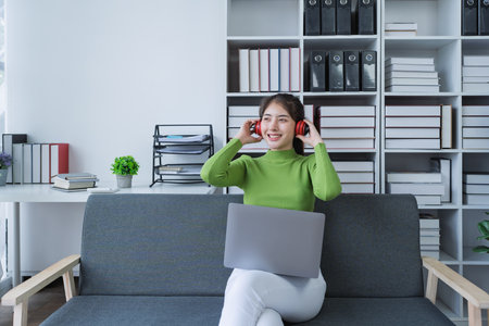 Woman sitting comfortably on a couch with headphones, relaxing to music while using a computer in a cozy setting.の写真素材
