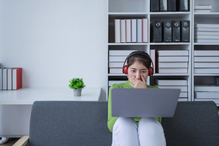 Woman sitting comfortably on a couch with headphones, relaxing to music while using a computer in a cozy setting.の写真素材