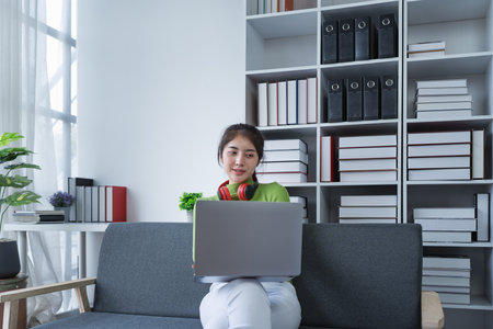 Woman sitting comfortably on a couch with headphones, relaxing to music while using a computer in a cozy setting.の写真素材