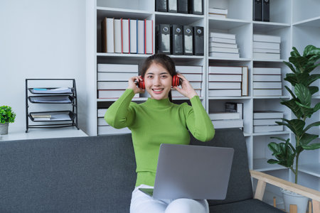 Woman sitting comfortably on a couch with headphones, relaxing to music while using a computer in a cozy setting.の写真素材
