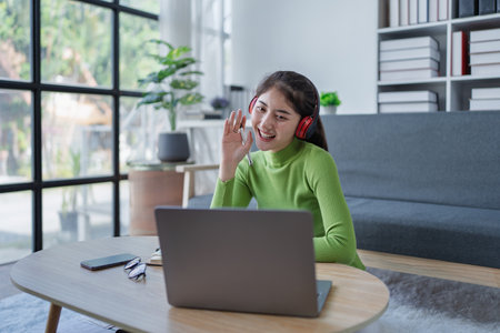 Young girl sitting on a couch with a laptop and headphones actively participating in a video conference call in a cozy home.の写真素材