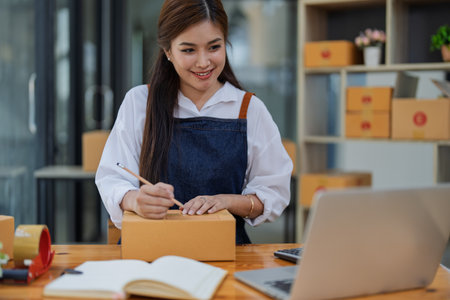 Young woman engaged in online business working with using laptop computer, preparing package for shipping and taking notes customer address at box.の写真素材