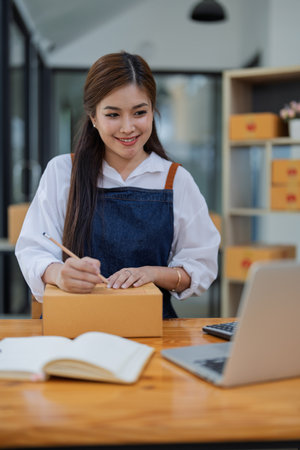 Young woman engaged in online business working with using laptop computer, preparing package for shipping and taking notes customer address at box.の写真素材