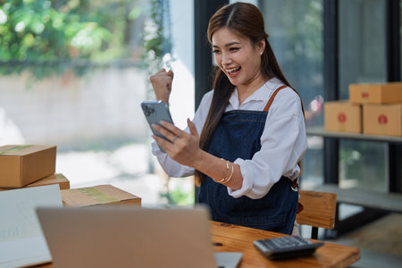 Excited young woman celebrating success while using smartphone to see data delivery products customer profit Selling products of This month.の写真素材