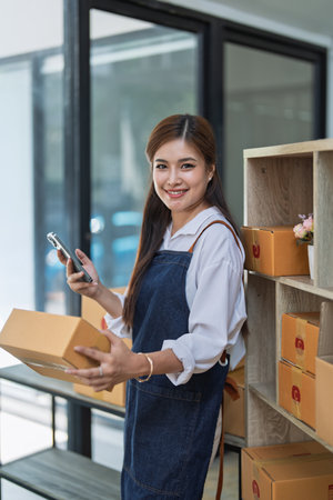 Young woman engaged in online business working with using smartphone, preparing package for shipping and taking notes customer address at box.の写真素材