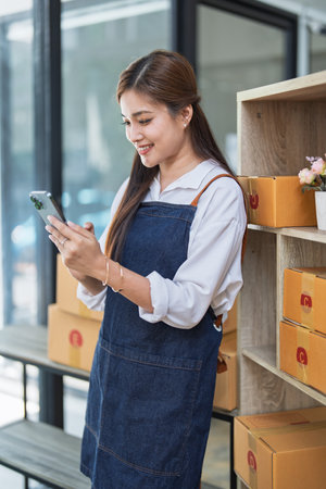 Young woman engaged in online business working with using smartphone, preparing package for shipping and taking notes customer address at box.の写真素材