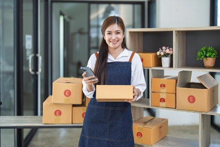 Young woman engaged in online business working with using smartphone, preparing package for shipping and taking notes customer address at box.の写真素材