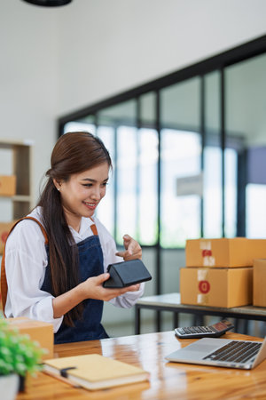 Young woman engaged in online business while packing delivery product at a wooden table while working on laptop for video to customer, Online selling and logistics concepts.の写真素材