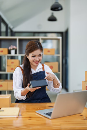 Young woman engaged in online shopping while unboxing a package at home office surrounded by cardboard boxes.の写真素材