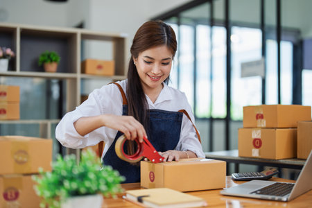 Young woman happily packing boxes with tape at a modern home office workspace surrounded by shipping supplies.の写真素材