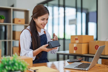 Young woman engaged in online business while packing delivery product at a wooden table while working on laptop for video to customer, Online selling and logistics concepts.の写真素材