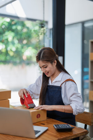 Young woman happily packing boxes with tape at a modern home office workspace surrounded by shipping supplies.の写真素材