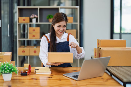 Young woman engaged in online business while packing delivery boxes at a wooden table while working on laptop for video to customer, Online selling and logistics concepts.の写真素材