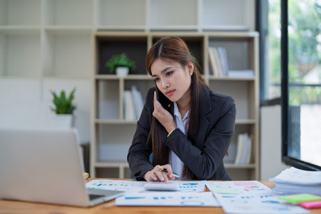 Professional businesswoman engaged in smartphone call by calculator to calculate finance with using stock marketing charts, notebook, and laptop in office.の写真素材
