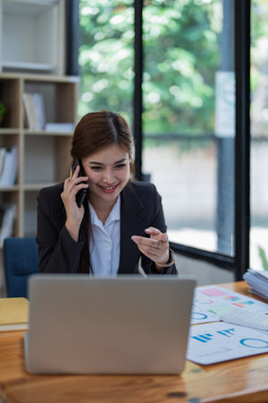Professional businesswoman engaged in smartphone call using calculator to calculate finance with stock marketing charts, notebook, and laptop in office.の写真素材