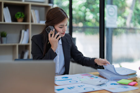Professional businesswoman engaged in smartphone call by calculator to calculate finance with using stock marketing charts, notebook, and laptop in office.の写真素材