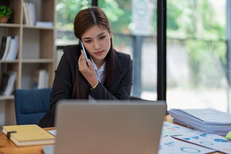 Professional businesswoman engaged in smartphone call by calculator to calculate finance with using stock marketing charts, notebook, and laptop in office.の写真素材