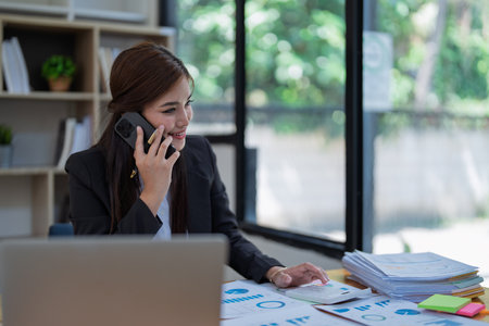 Professional businesswoman engaged in smartphone call by calculator to calculate finance with using stock marketing charts, notebook, and laptop in office.の写真素材