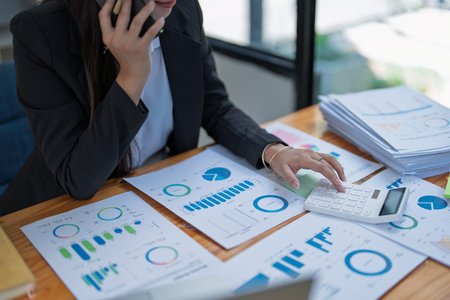 Professional businesswoman engaged in smartphone call by calculator to calculate finance with using stock marketing charts, notebook, and laptop in office.の写真素材