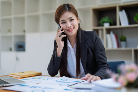 Professional businesswoman engaged in smartphone call by calculator to calculate finance with using stock marketing charts, notebook, and laptop in office.の写真素材