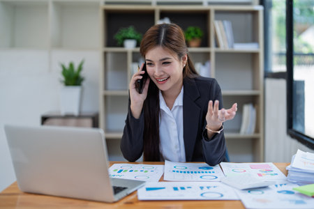 Professional businesswoman engaged in smartphone call by calculator to calculate finance with using stock marketing charts, notebook, and laptop in office.の写真素材