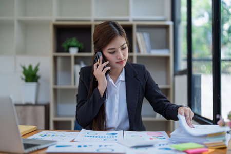 Professional businesswoman engaged in smartphone call by calculator to calculate finance with using stock marketing charts, notebook, and laptop in office.の写真素材