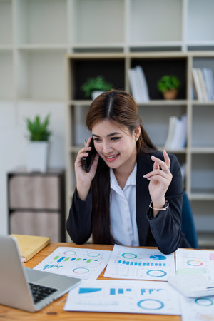Professional businesswoman engaged in smartphone call by calculator to calculate finance with using stock marketing charts, notebook, and laptop in office.の写真素材