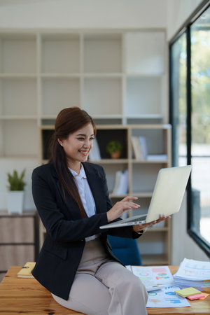 Smiling businesswoman in an office, working on a laptop while by financial, market charts documents and showcasing video conference professional engagement.の写真素材