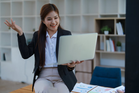 Smiling businesswoman in an office, working on a laptop while by financial, market charts documents and showcasing video conference professional engagement.の写真素材