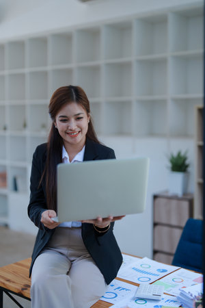 Smiling businesswoman in an office, working on a laptop while by financial, market charts documents and showcasing video conference professional engagement.の写真素材