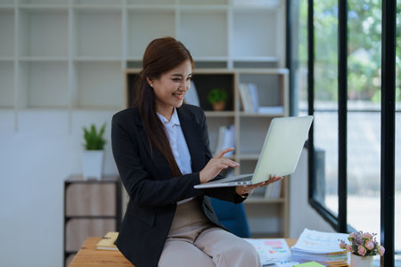 Smiling businesswoman in an office, working on a laptop while by financial, market charts documents and showcasing video conference professional engagement.の写真素材