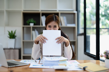 Young smiling woman playfully hiding behind large blank paper sheet at modern office desk with documents stacked in front of her for creative concept.の写真素材