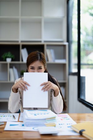 Young woman in beige long-sleeved shirt playfully hiding her face with a blank sheet at a desk cluttered with heaps of paperwork and documents..の写真素材