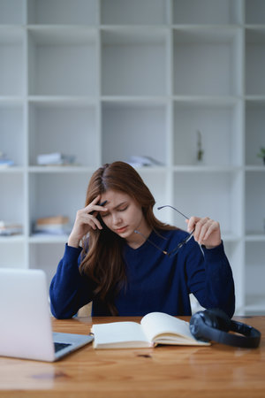 Stressed young woman overwhelmed by e-learning studies at desk with laptop, notebook, headphones, hands clasped to her face in frustration.の写真素材