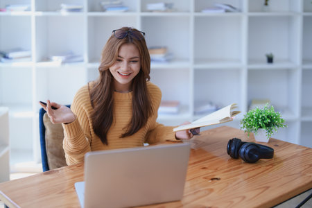 Smiling young woman engaged in a joyful online e-learning video call, gesturing while holding a book with headphones on a wooden table nearby.の写真素材